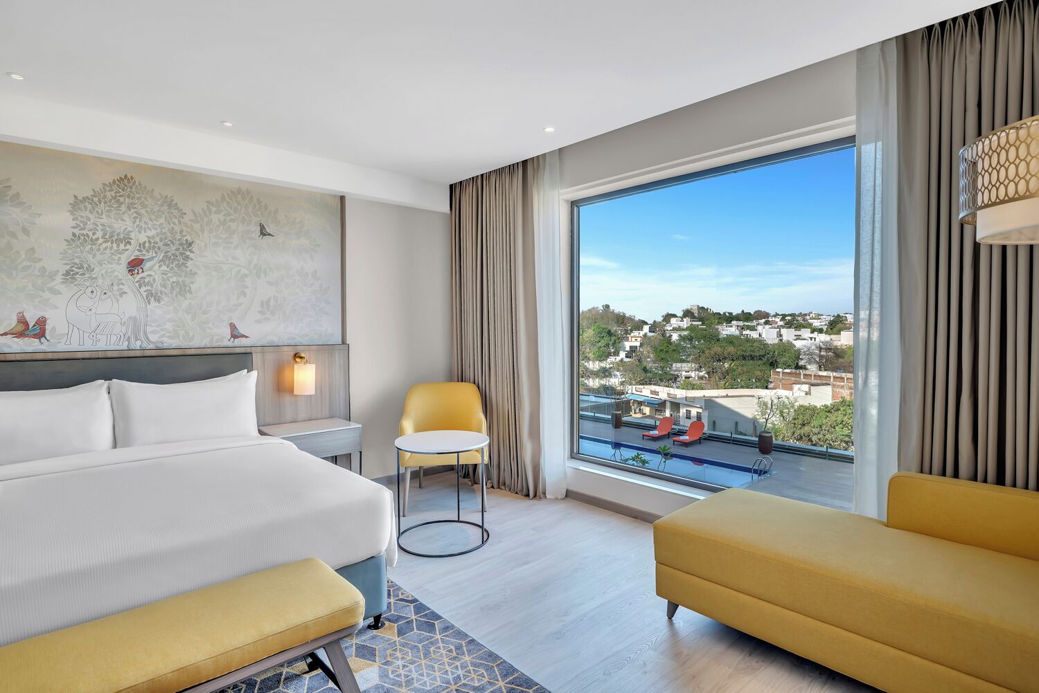 A hotel room with a large bed, a desk, a chair, a sofa, and a large window with a view of the city.