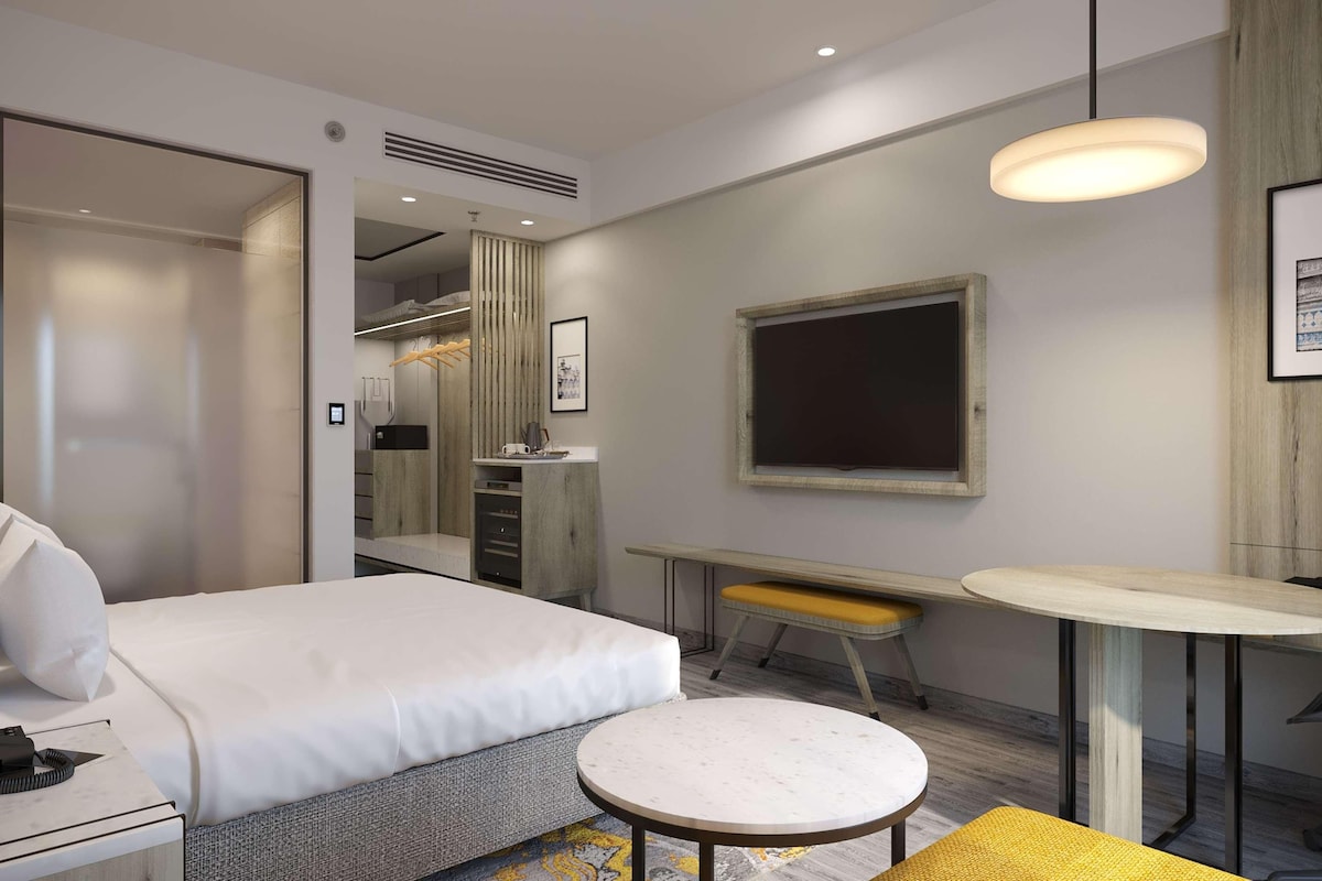 A modern hotel room with a bed, a desk, a chair, a small round table, and a large mirror.
