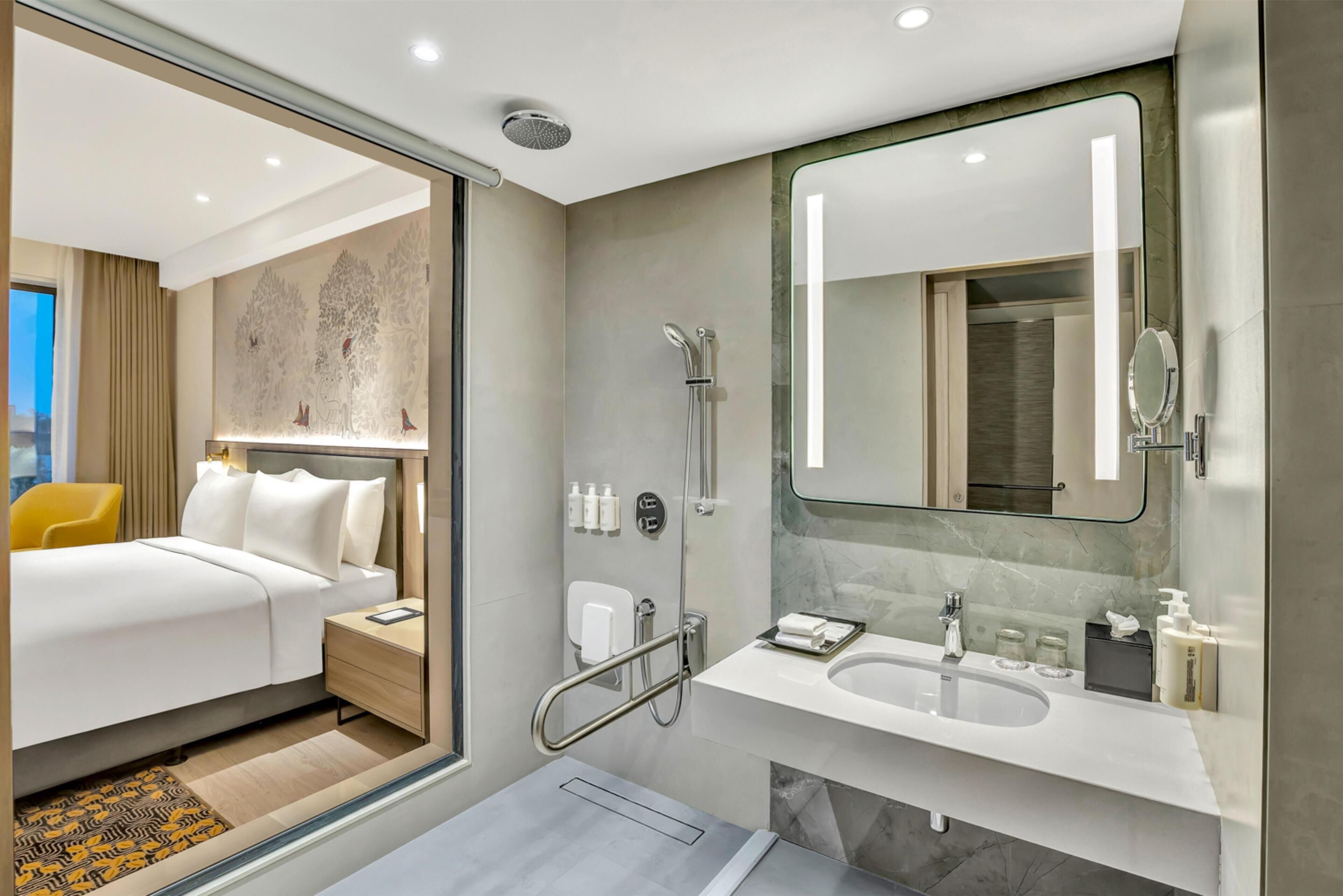 A modern hotel room with a bed, bedside table, and a bathroom with a shower, sink, and mirror.