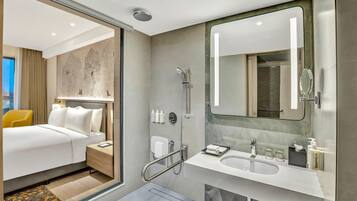 A modern hotel room with a bed, bedside table, and a bathroom with a shower, sink, and mirror.