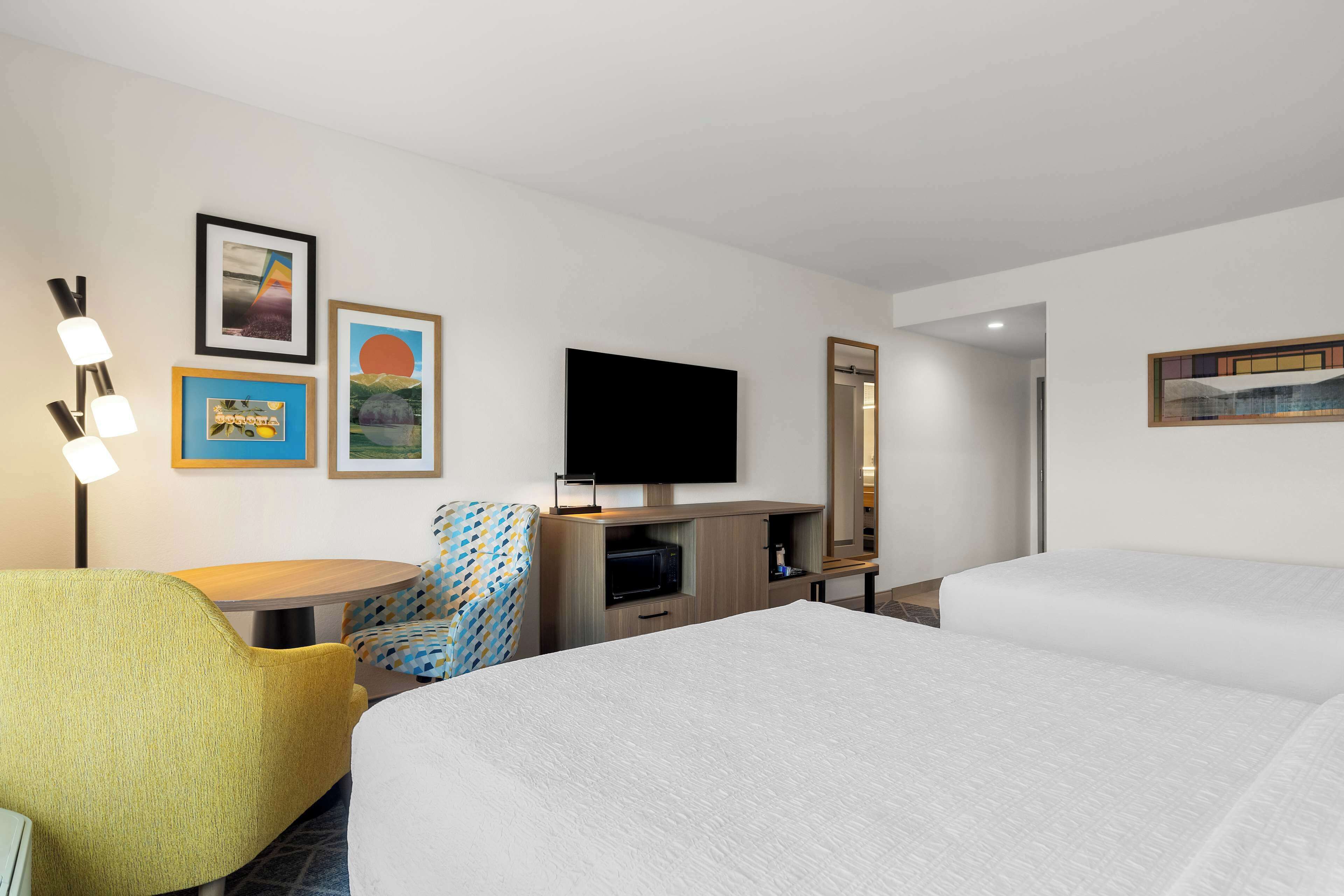Ein Hotelzimmer mit zwei Betten, einem Fernseher, einer Mikrowelle, einem Sessel und gerahmten Bildern an der Wand.