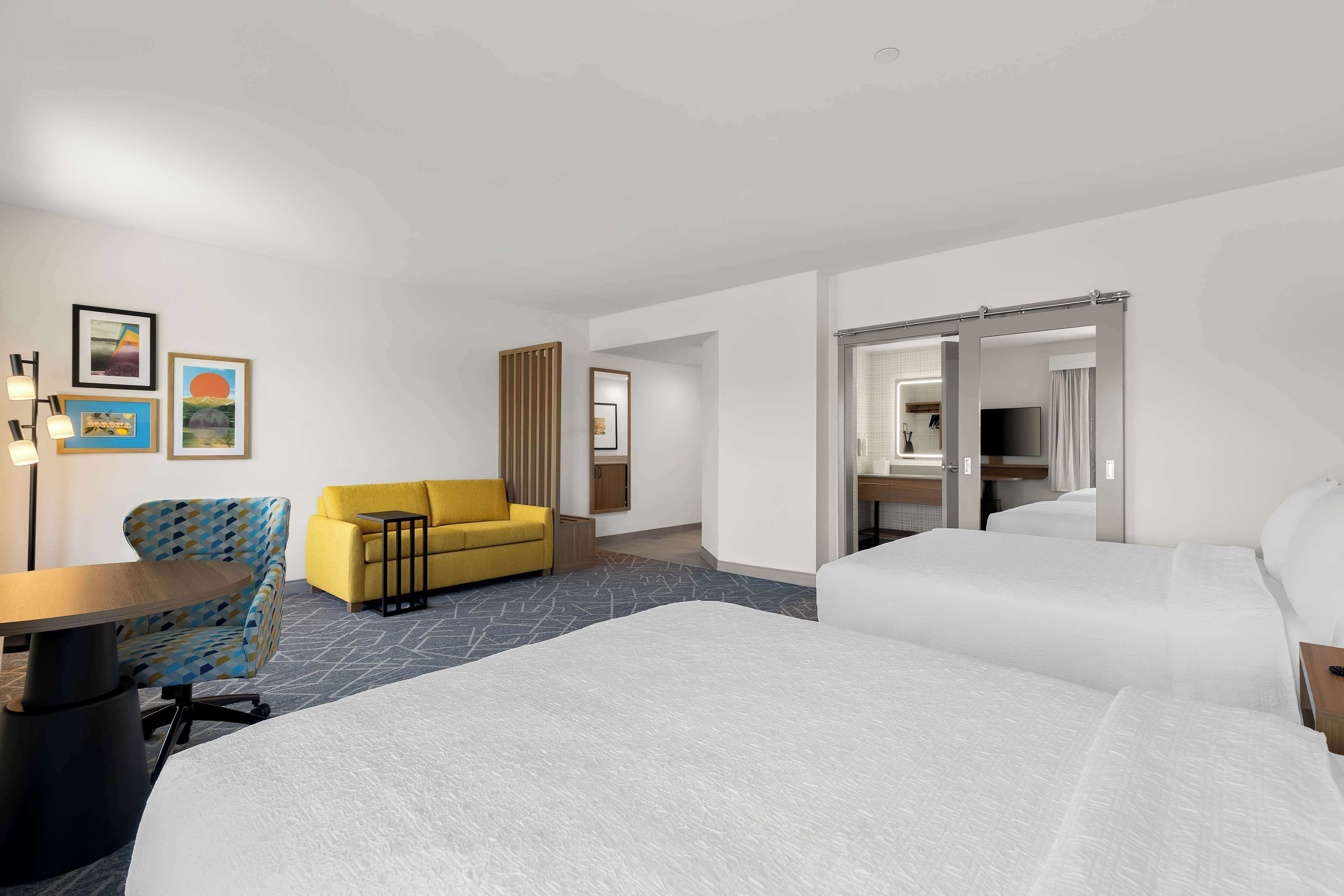 Habitación de hotel con una cama grande, un sofá, un escritorio y una silla. Hay cuadros en la pared y una puerta que da al baño.