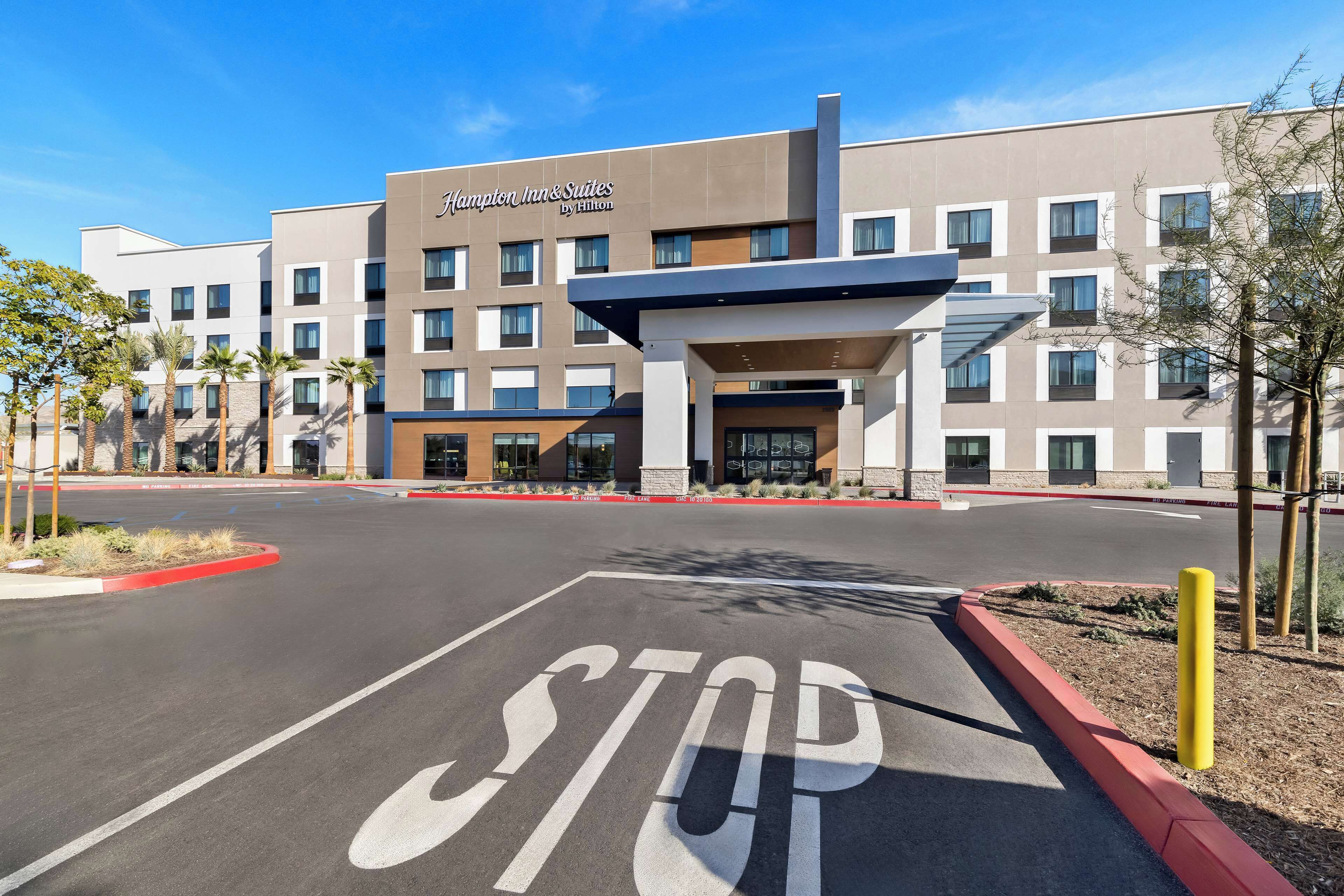 L’entrée d’un hôtel, avec un panneau « STOP » bien visible au premier plan, un ciel dégagé et un bâtiment doté de nombreuses fenêtres et d’une enseigne indiquant « Hampton Inn & Suites by Hilton ».
