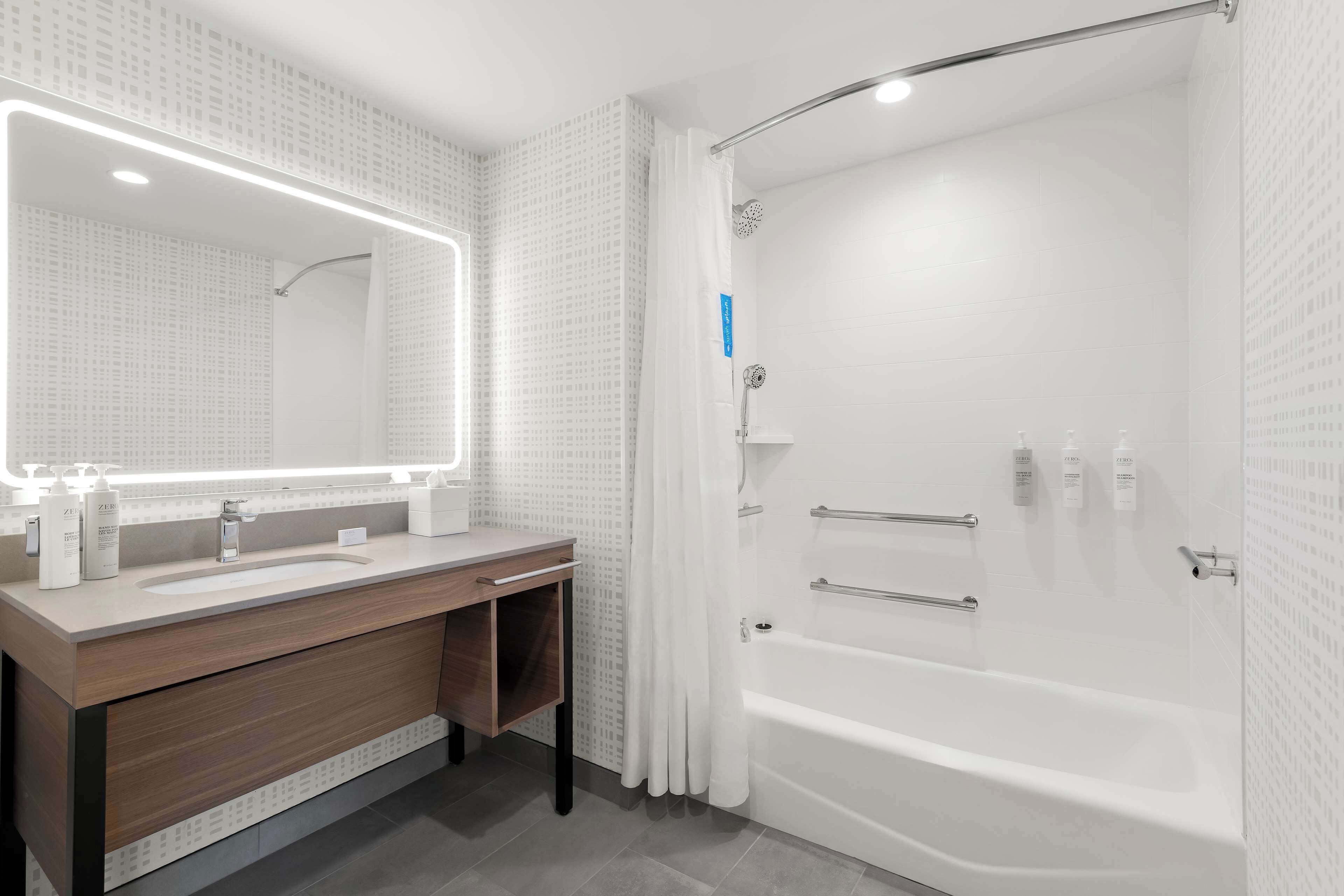 A modern bathroom with a large mirror, two sinks, a vanity, a bathtub, and a shower.
