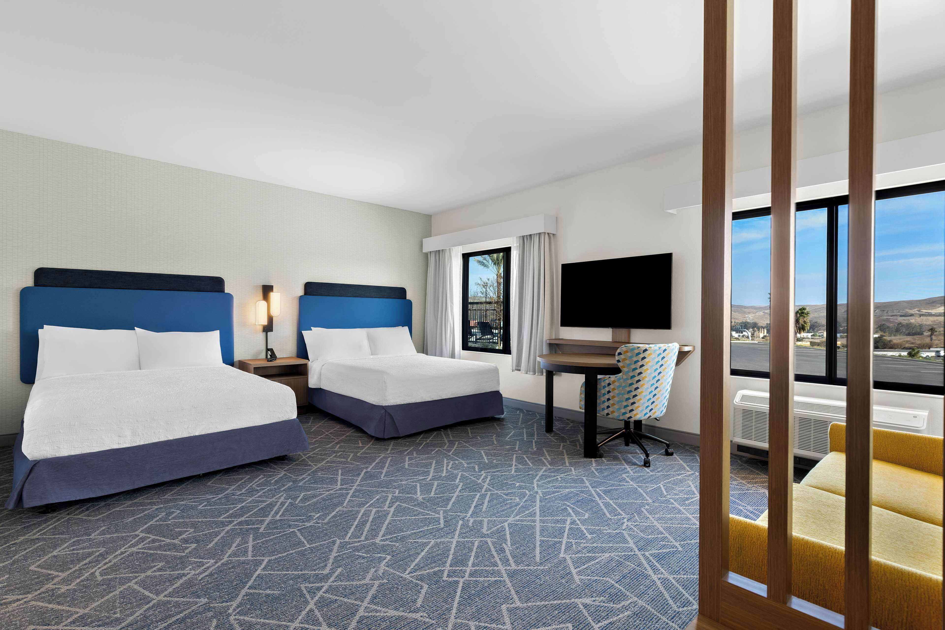 A hotel room with two beds, a desk, a TV, and a large window with a city view.