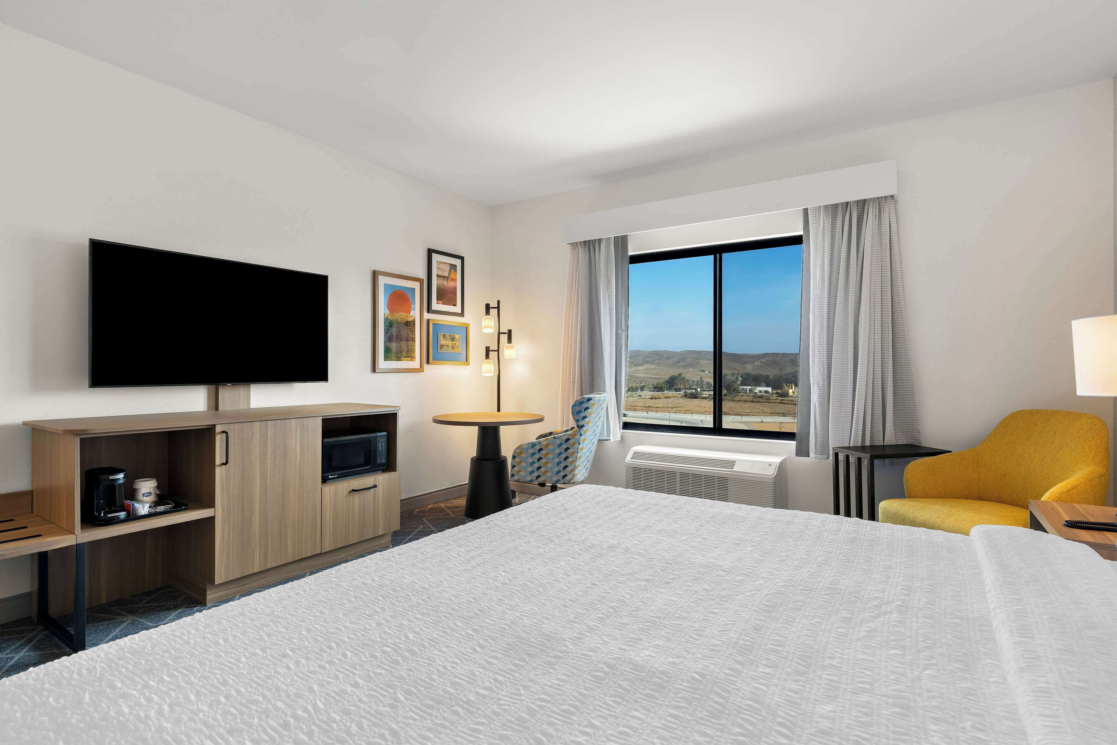 A hotel room with a large bed, a flat-screen TV, a small table, a chair, and a window with a view of the countryside.