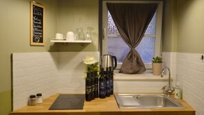 Une cuisine compacte avec un plan de travail en bois, un évier en acier inoxydable et une fenêtre avec un rideau. On y trouve des salières et des poivrières, un vase avec une fleur, ainsi qu’une étagère avec des tasses et une bouilloire.