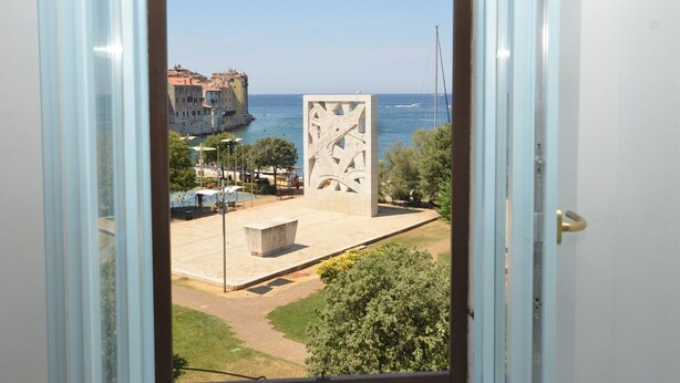 A view through a window of a coastal area with a large sculpture, a beach, and historic buildings. - Sunny Rovinj Apartment with Sea View - One Bedroom Apartment with Sea View (Rovinj)