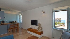 A modern living room with a sofa, a wooden coffee table, and a stone sculpture. There is a kitchen area with a microwave, a kettle, and a sink. A TV is mounted on the wall, and a window offers a view of a coastal town. - Sunny Rovinj Apartment with Sea View - One Bedroom Apartment with Sea View (Rovinj)