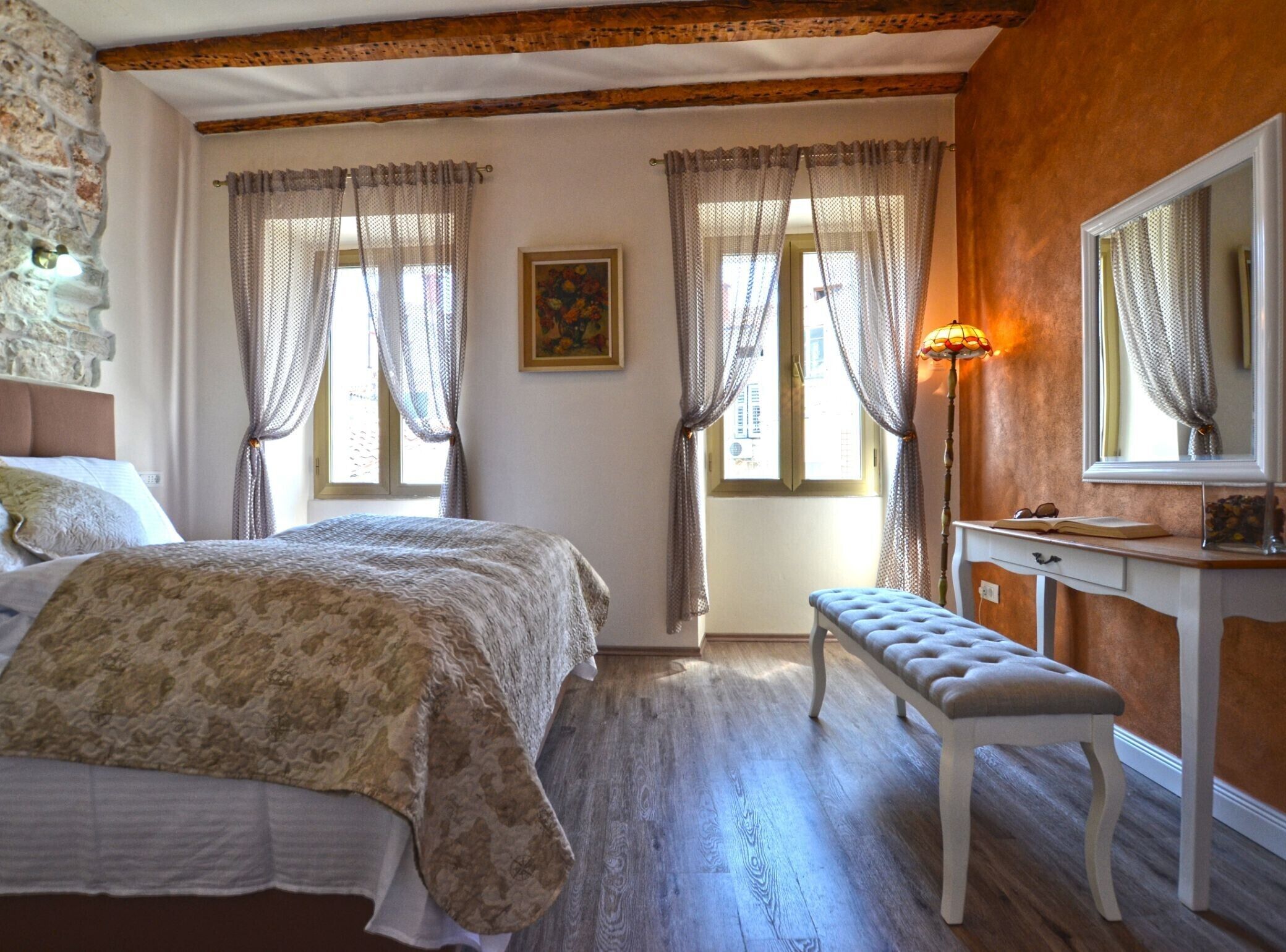 Quarto com cama, um banquinho, cômoda e duas janelas com cortinas.
