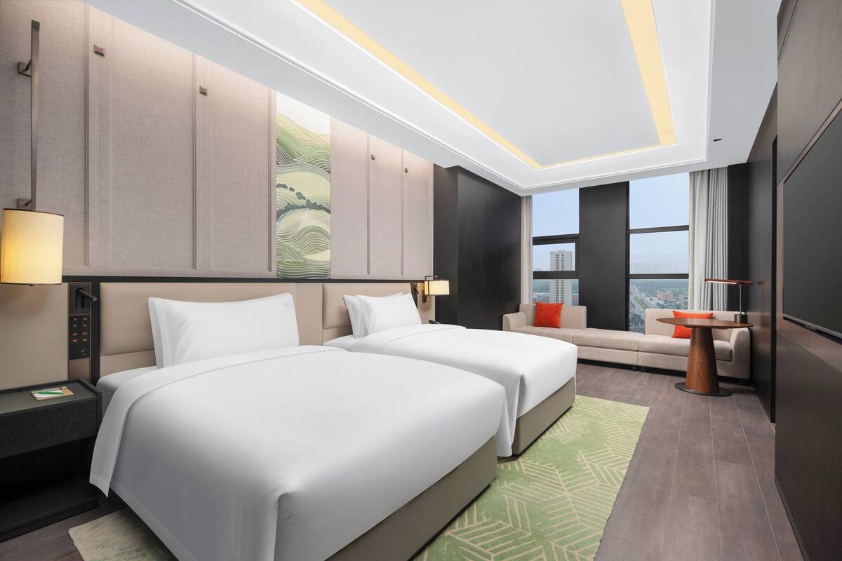 A modern hotel room with two beds, a sofa, a small table, and a large window offering a city view.
