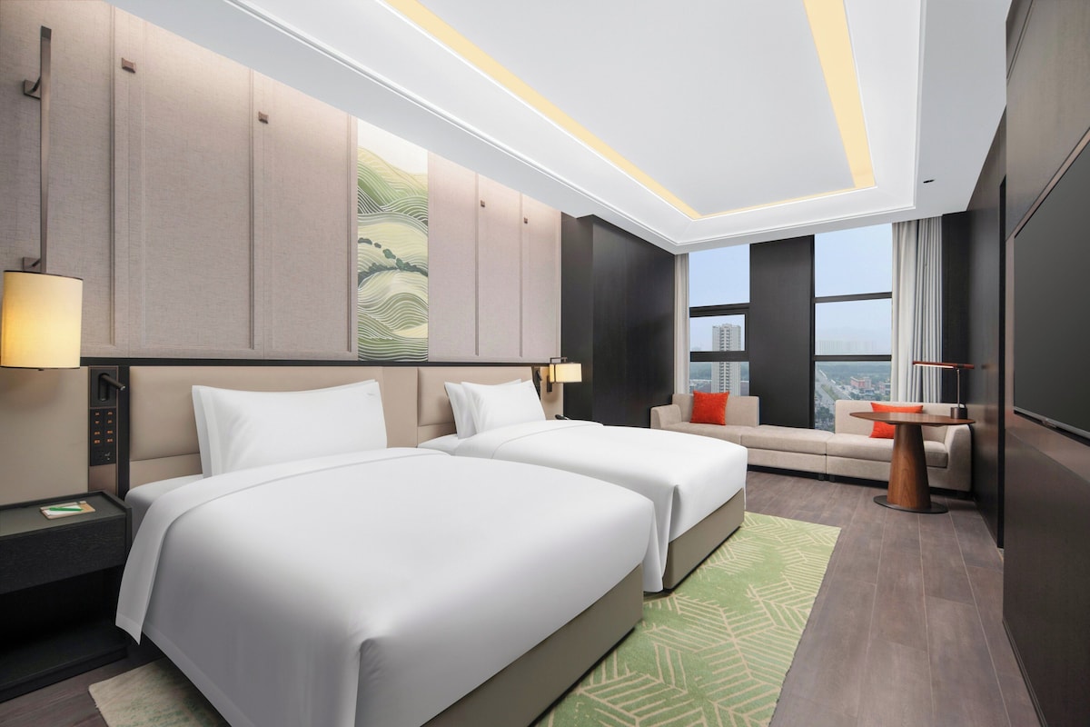 A modern hotel room with two beds, a sofa, a small table, and a large window offering a city view.