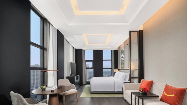Ein modernes Hotelzimmer mit einem großen Bett, einer Sitzecke, einem Esstisch und einem Fernseher. Das Zimmer verfügt über große Fenster und Blick auf die Stadt.