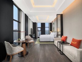 Habitación de hotel moderna con una cama grande, un sofá, una mesa de comedor y un televisor. La habitación tiene ventanas amplias y vistas a la ciudad.