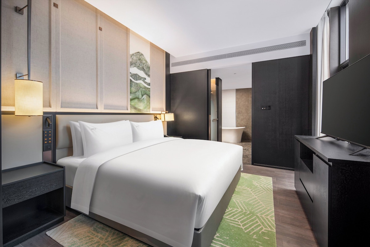 A modern hotel room with a large bed, bedside tables, a flat-screen TV, and a painting on the wall.