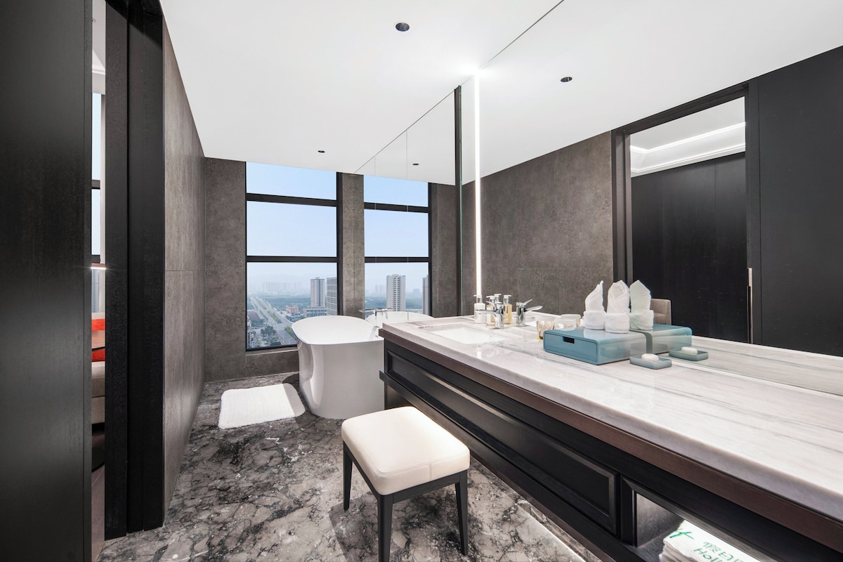 A modern bathroom with a large mirror, two sinks, a bathtub, and a city view.