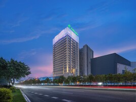Un hotel moderno de gran altura con un letrero verde llamativo en la parte superior, rodeado de árboles y una calle bien iluminada al atardecer.