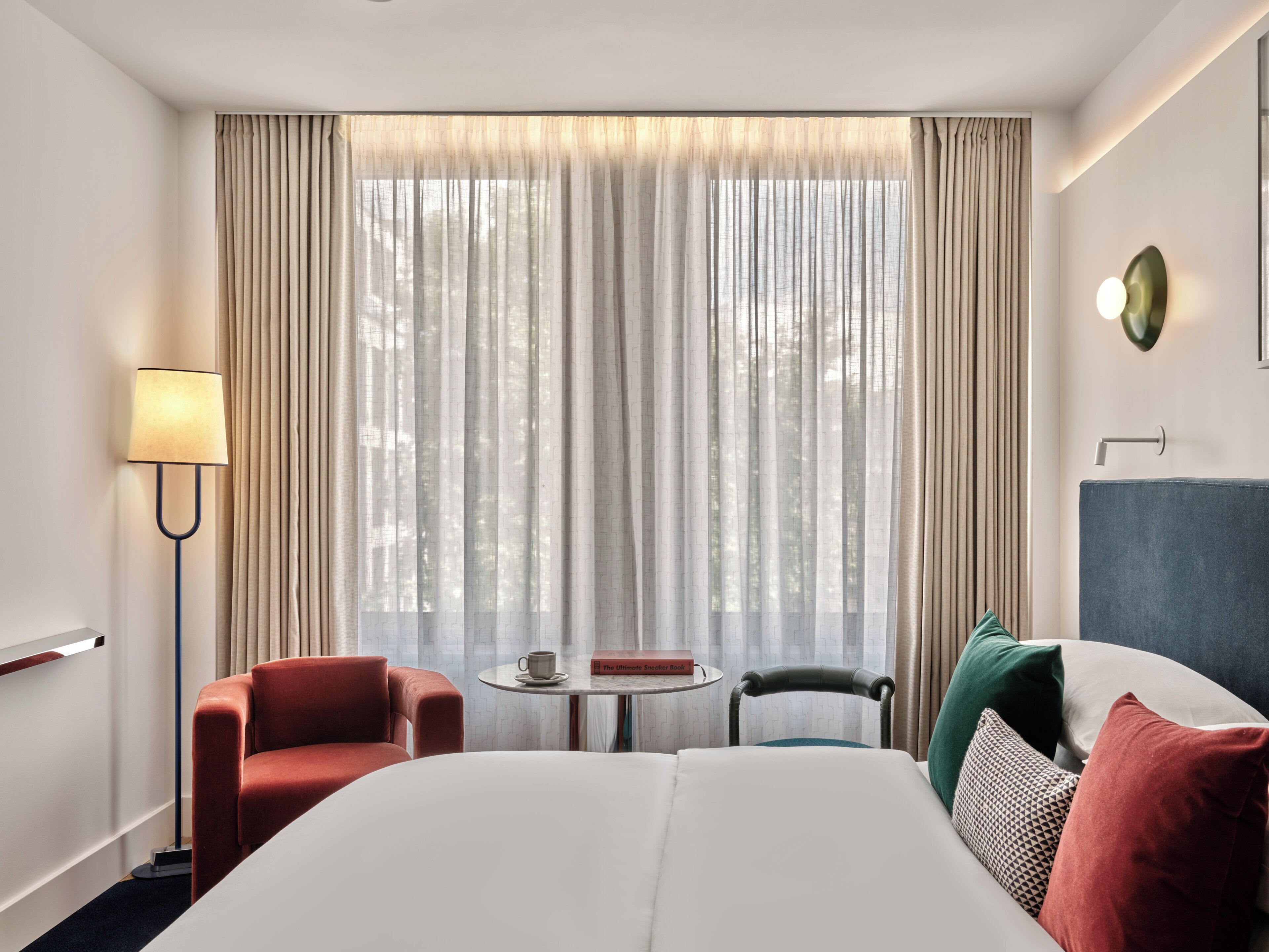 A hotel room with a bed, two chairs, a small table with a book and a cup, and a lamp.