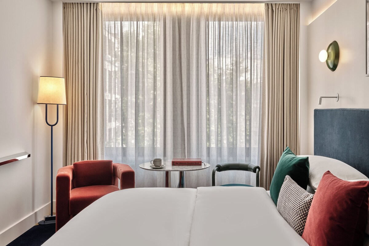 Une chambre d’hôtel avec un lit, deux chaises, une petite table sur laquelle se trouvent un livre et une tasse, et une lampe.