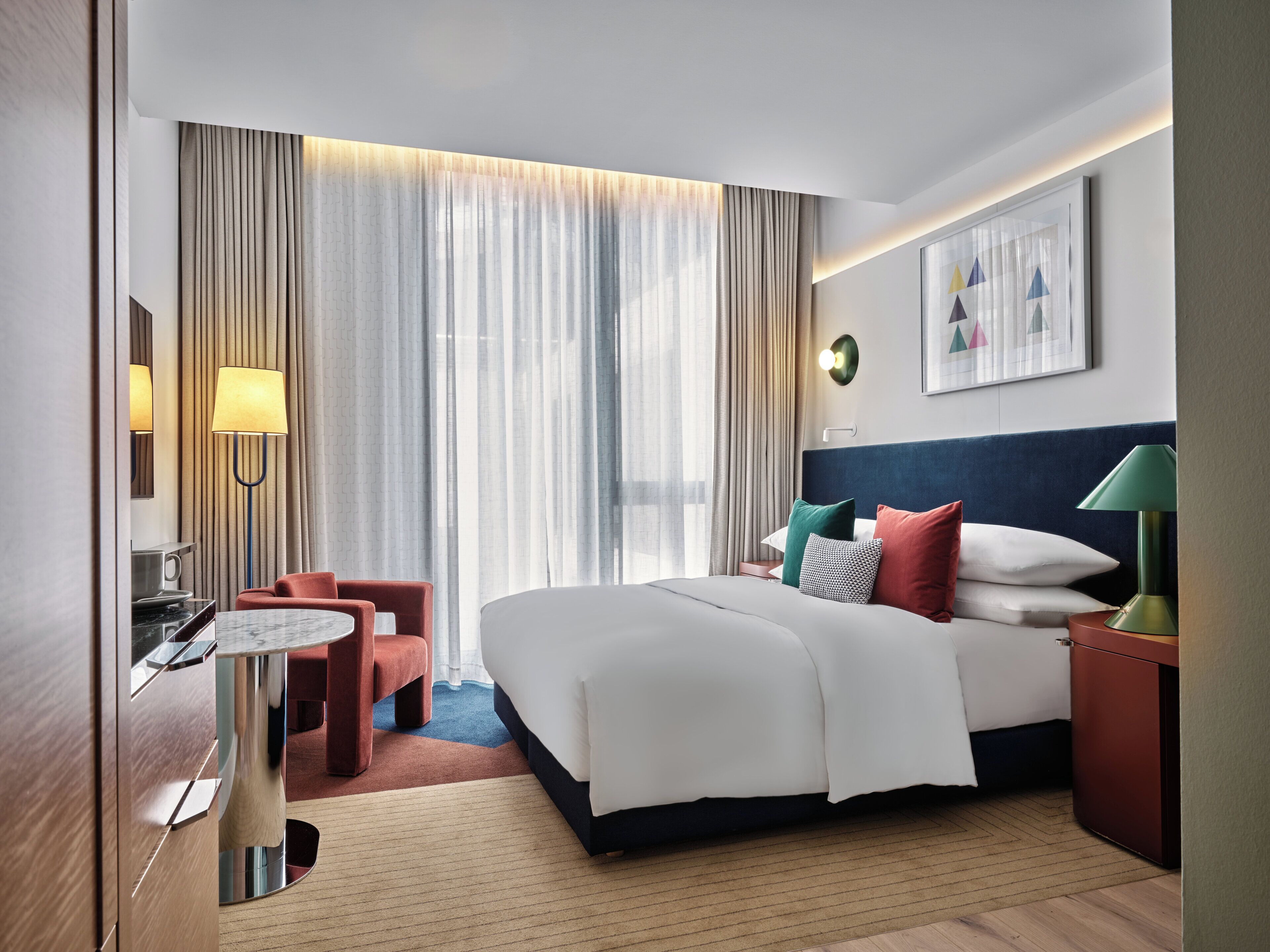 A hotel room with a bed, bedside tables, a desk, a chair, and a large window with curtains.