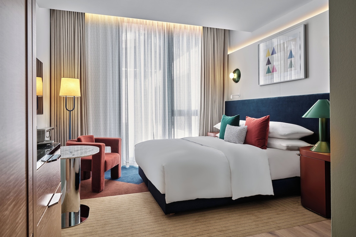 Une chambre d’hôtel comprenant un lit, des tables de chevet, un bureau, une chaise et une grande fenêtre avec des rideaux.