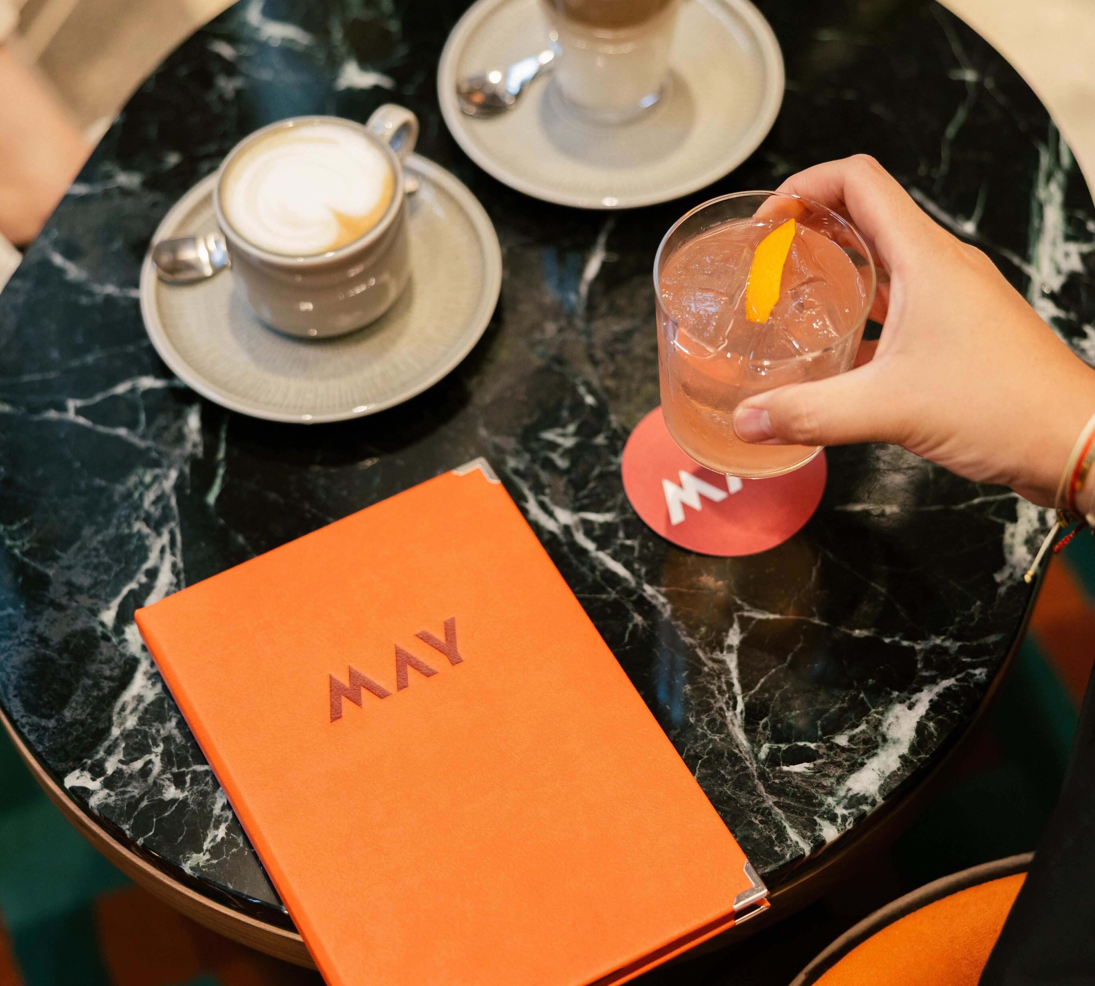 A table with a coffee cup, a glass of iced drink, and a book with the word 'MAY' on it.