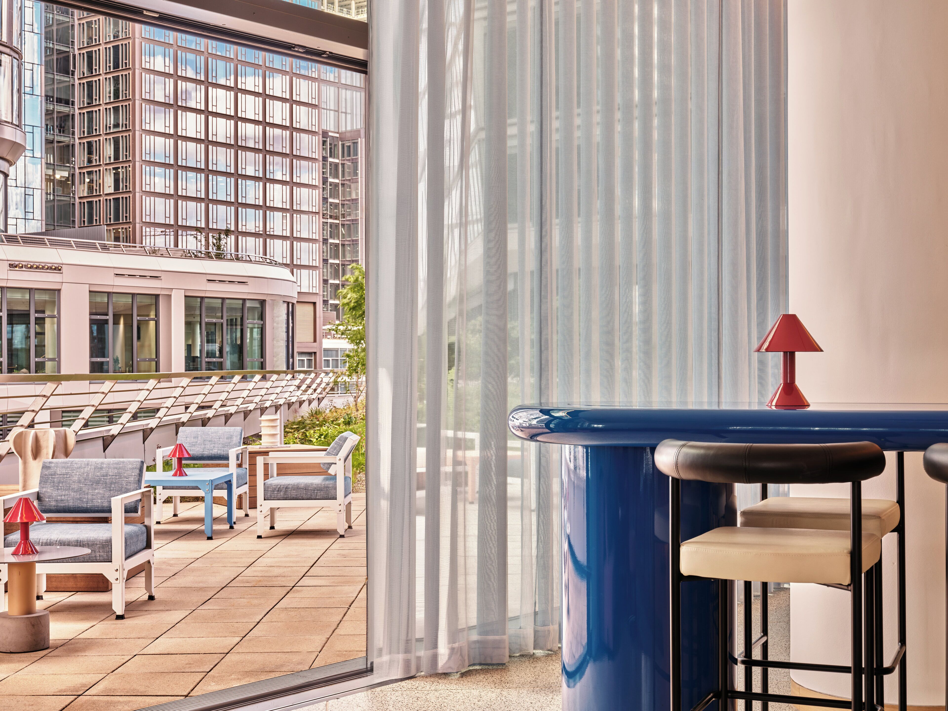 Ein Balkon mit Sitzgelegenheit im Freien, Blick auf ein Gebäude und eine rote Lampe auf einem Tisch.