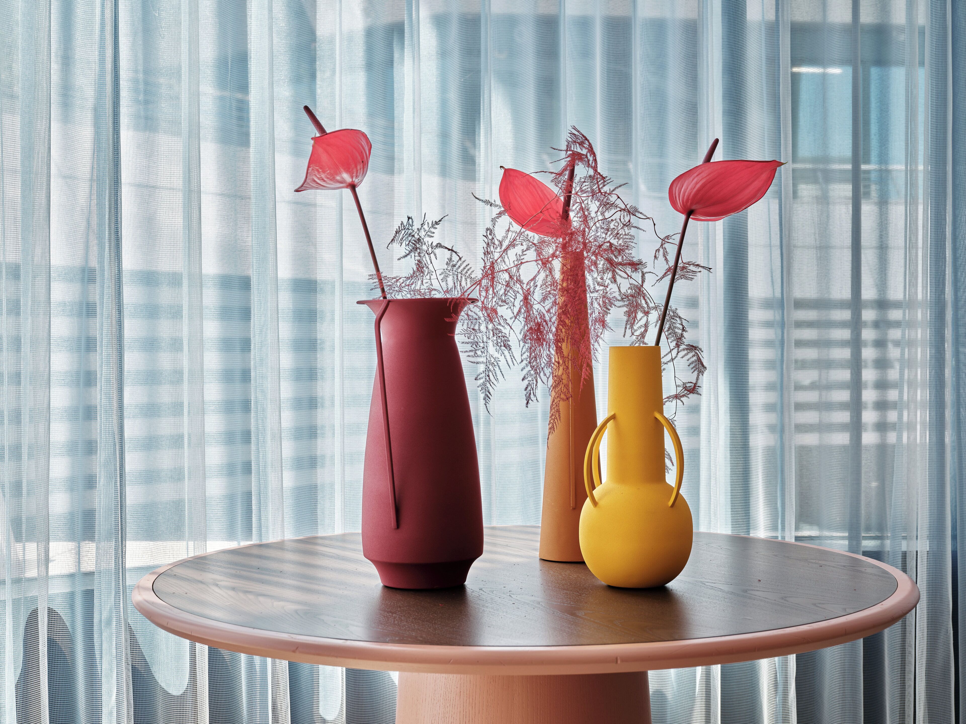 A table with three vases, each with a different shape and color, placed on a round table against a window with sheer curtains.
