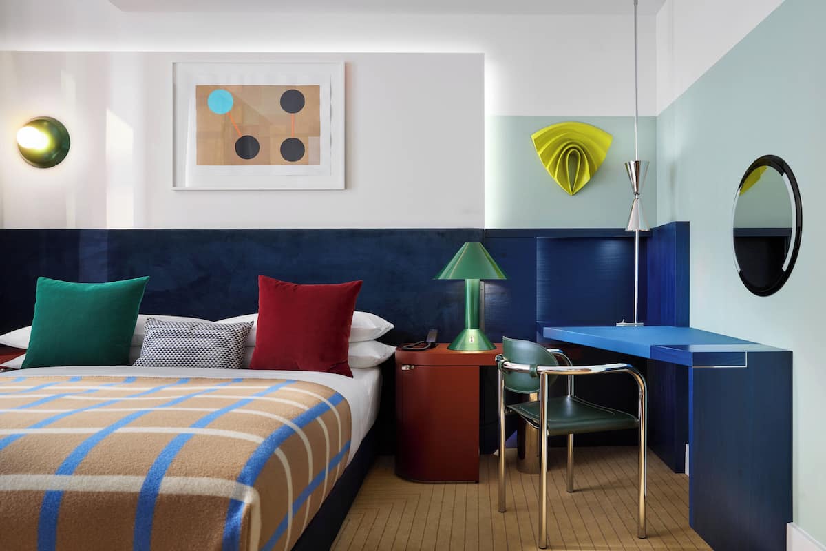 Ein Hotelzimmer mit Bett, Schreibtisch, Stuhl, Lampe, Spiegel und einem Wandbild.