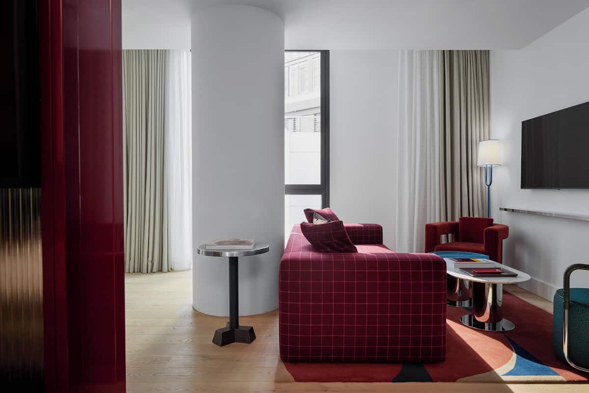 Ein modernes Hotelzimmer mit einem roten Sofa, einem kleinen runden Tisch, einem an der Wand montierten Fernseher und einem Fenster mit Vorhängen.
