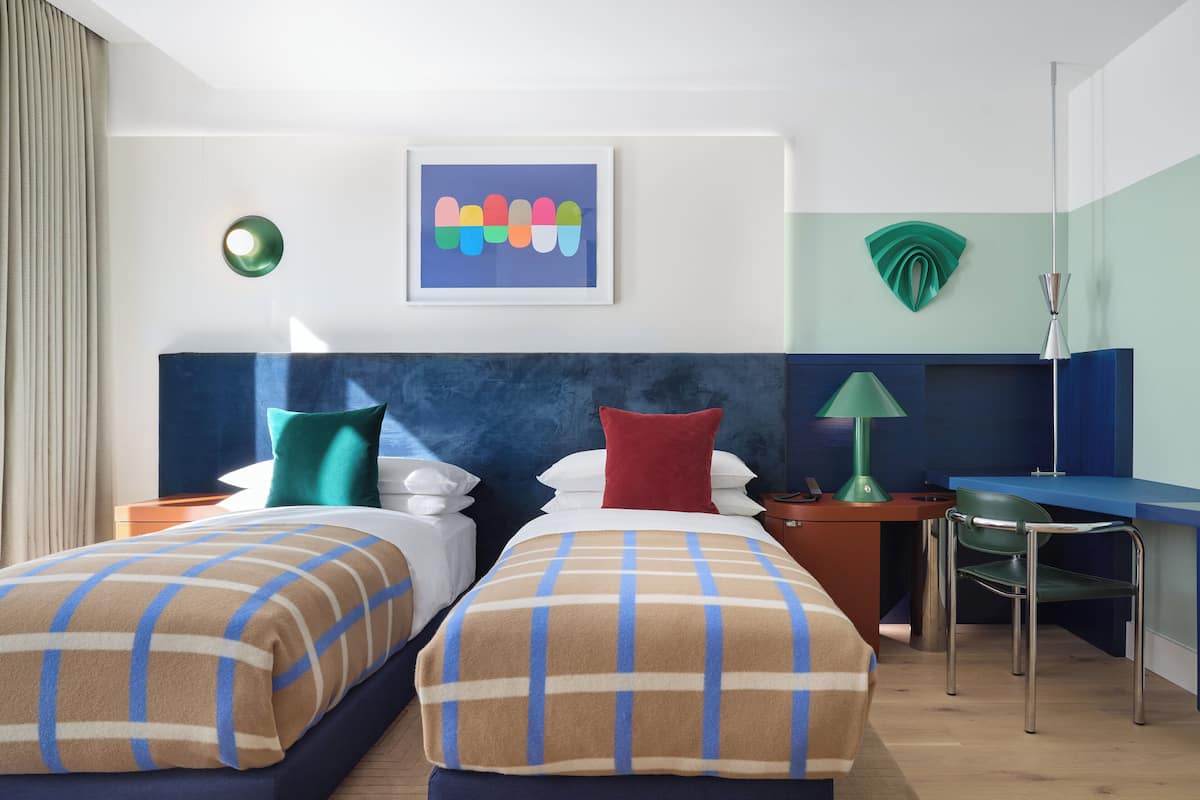 Ein Hotelzimmer mit zwei Betten, einem Schreibtisch, einem Stuhl, einer Lampe, einem Bild und einer Wanddekoration.