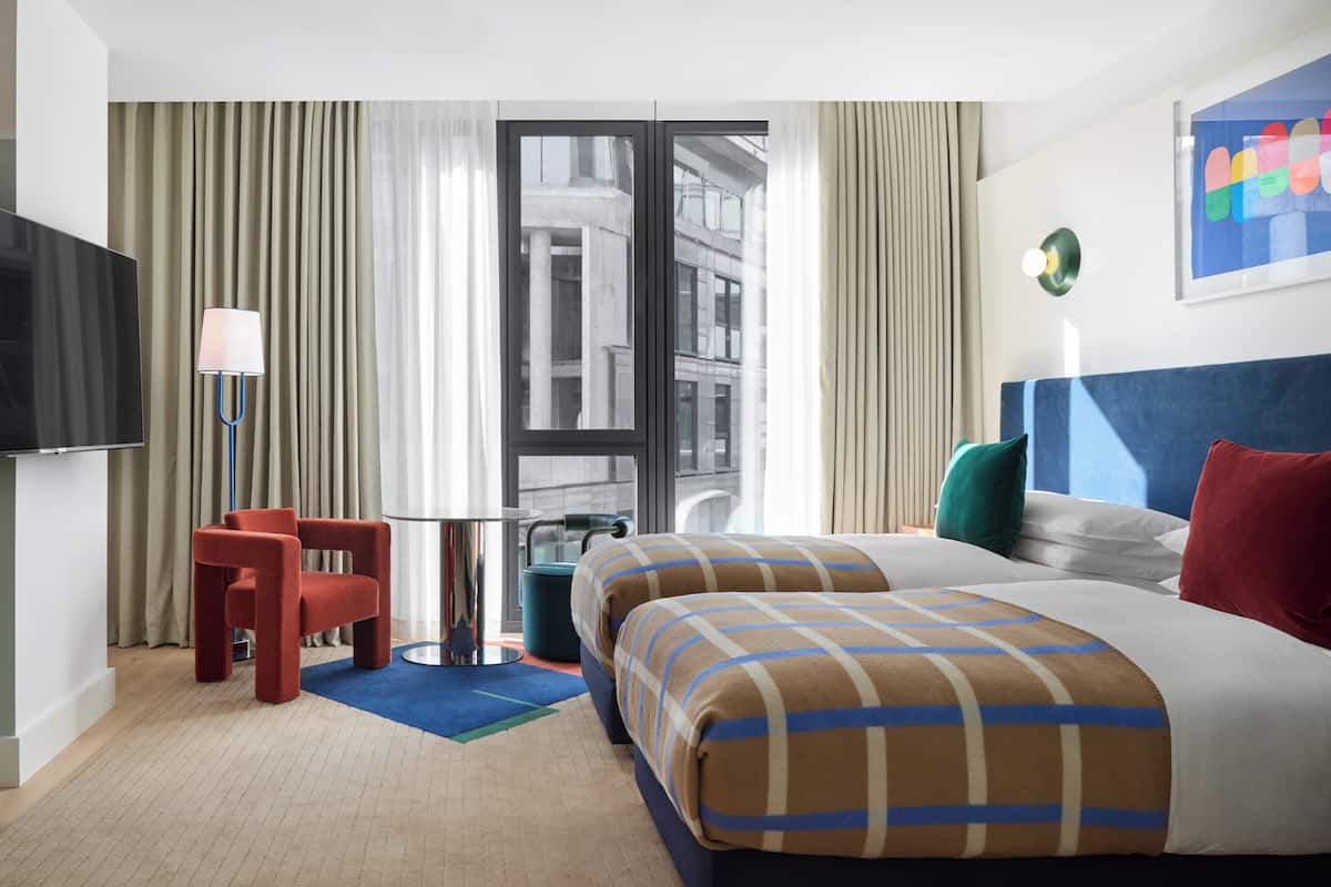 Ein Hotelzimmer mit einem Bett, zwei Kissen, einem Fernseher, einem Sessel und einem Fenster mit Vorhängen.