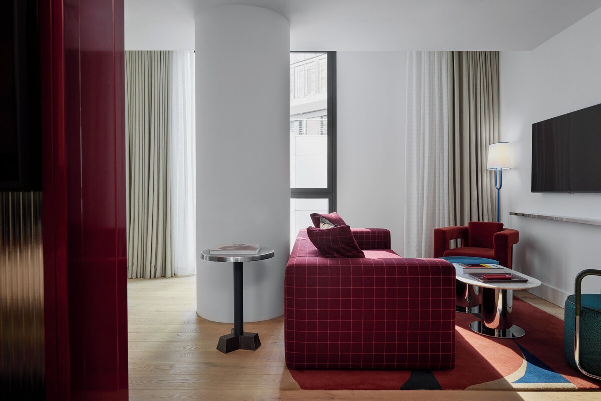 Une chambre d’hôtel moderne avec un canapé rouge, une petite table ronde, un téléviseur fixé au mur et une fenêtre avec des rideaux.