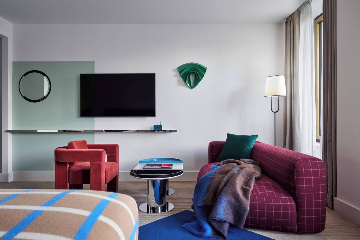 Ein modernes Hotelzimmer mit Flachbildfernseher, einem runden Spiegel, einem kleinen Tisch mit Lampe, einem roten Sofa und einer blauen Decke.