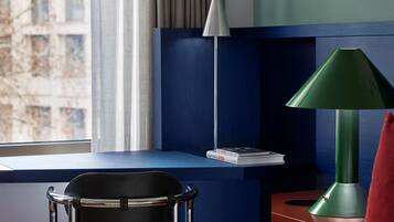 A hotel room with a bed, a desk, a chair, a green lamp, and a view of buildings outside the window.