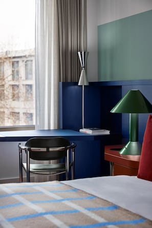 Une chambre d’hôtel avec un lit, un bureau, une chaise, une lampe verte et une vue sur les bâtiments à travers la fenêtre.