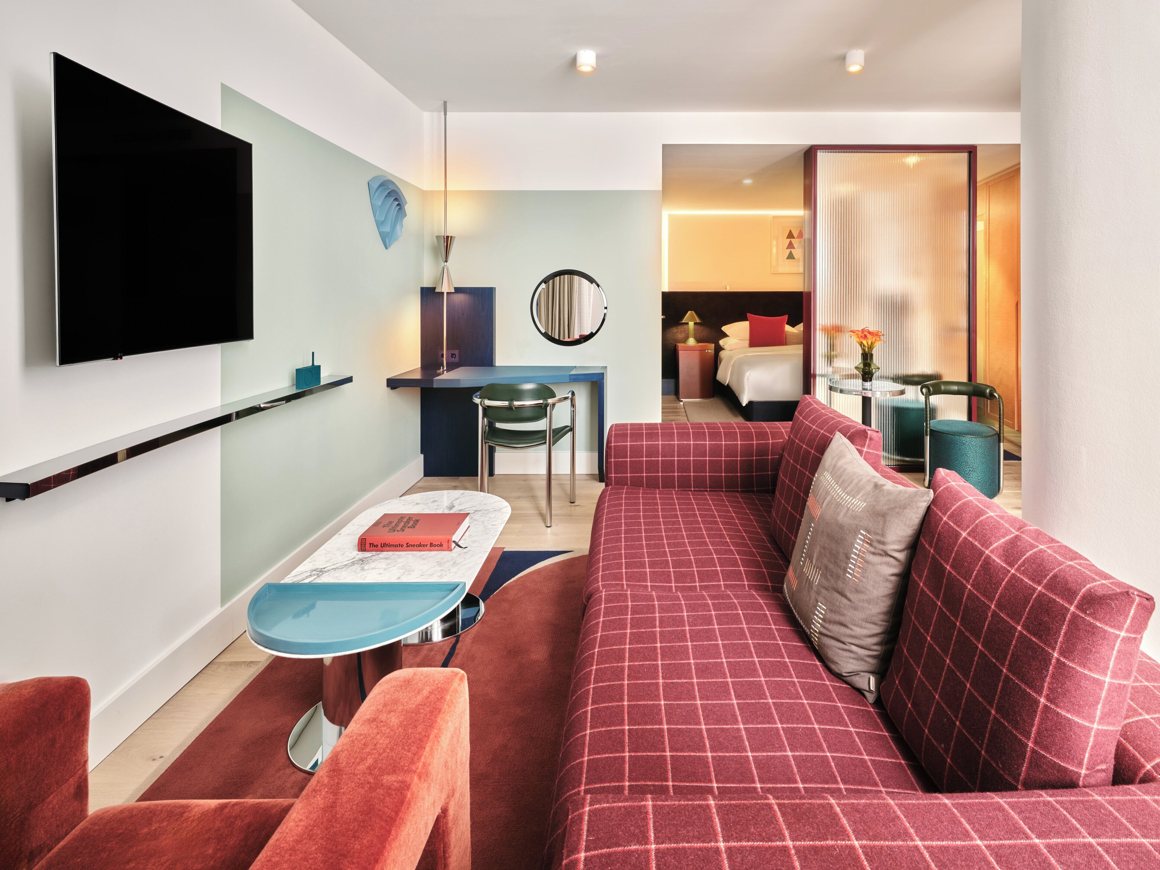 A modern hotel room with a flat-screen TV, a sofa, a dining table, and a bed with a red pillow.