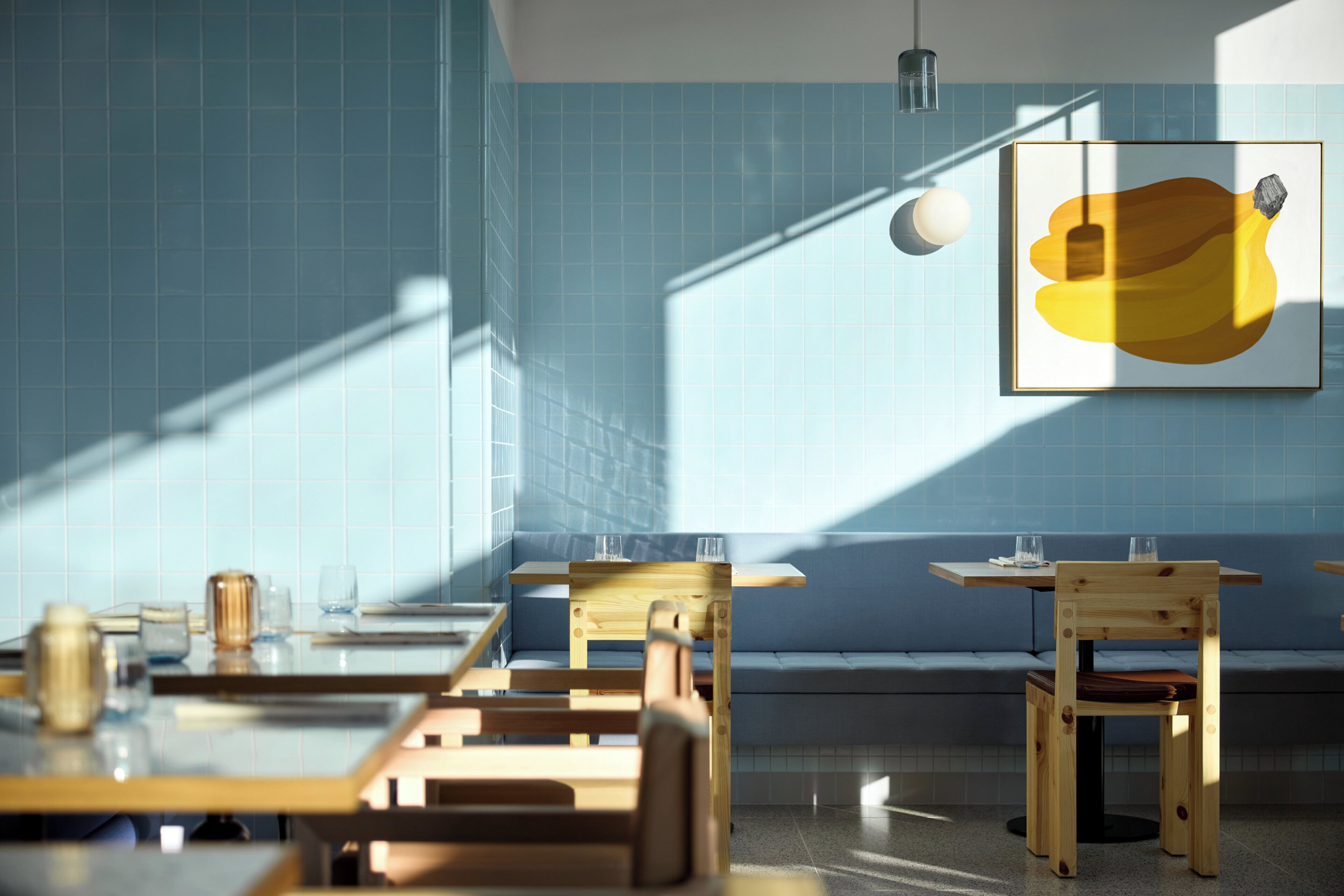 Ein modernes Restaurant-Interieur mit blauen Fliesenwänden, Holztischen und -stühlen sowie einem großen gerahmten Kunstwerk an der Wand.
