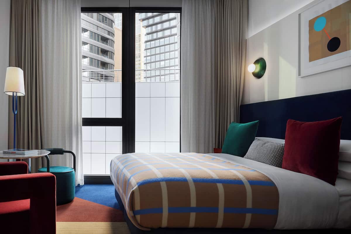 Ein Hotelzimmer mit einem großen Bett, einem Schreibtisch mit Lampe, einem Sessel und Blick auf die Stadt.