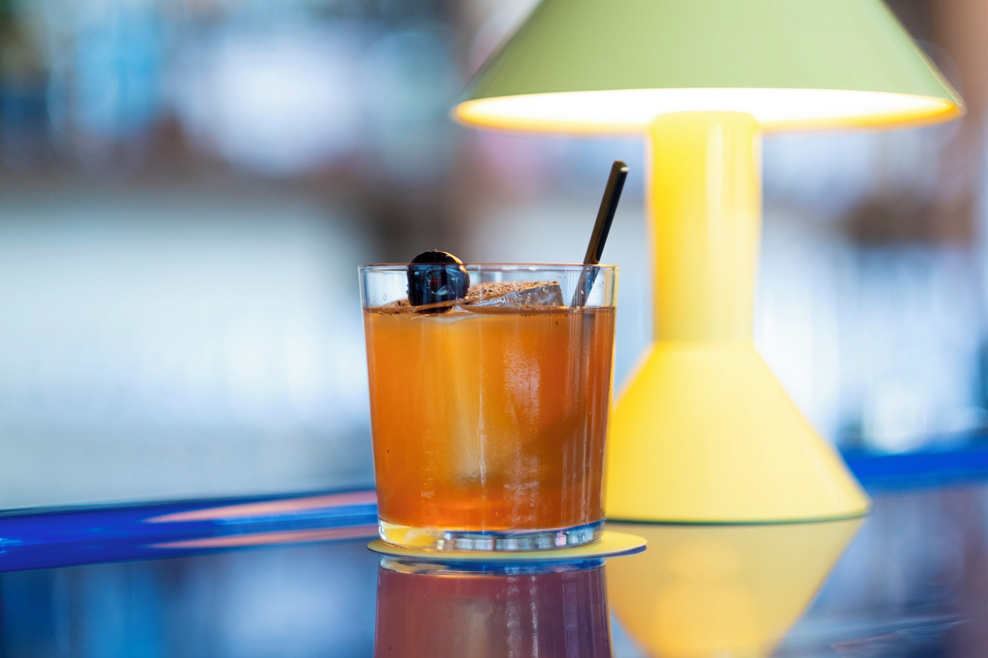 A glass of amber-colored beverage with a dark sphere and a straw, placed on a reflective surface with a yellow lamp in the background.
