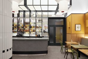 Un espace bar avec un comptoir à carreaux, des étagères remplies de bouteilles et un coin salon avec des tables et des chaises en bois.