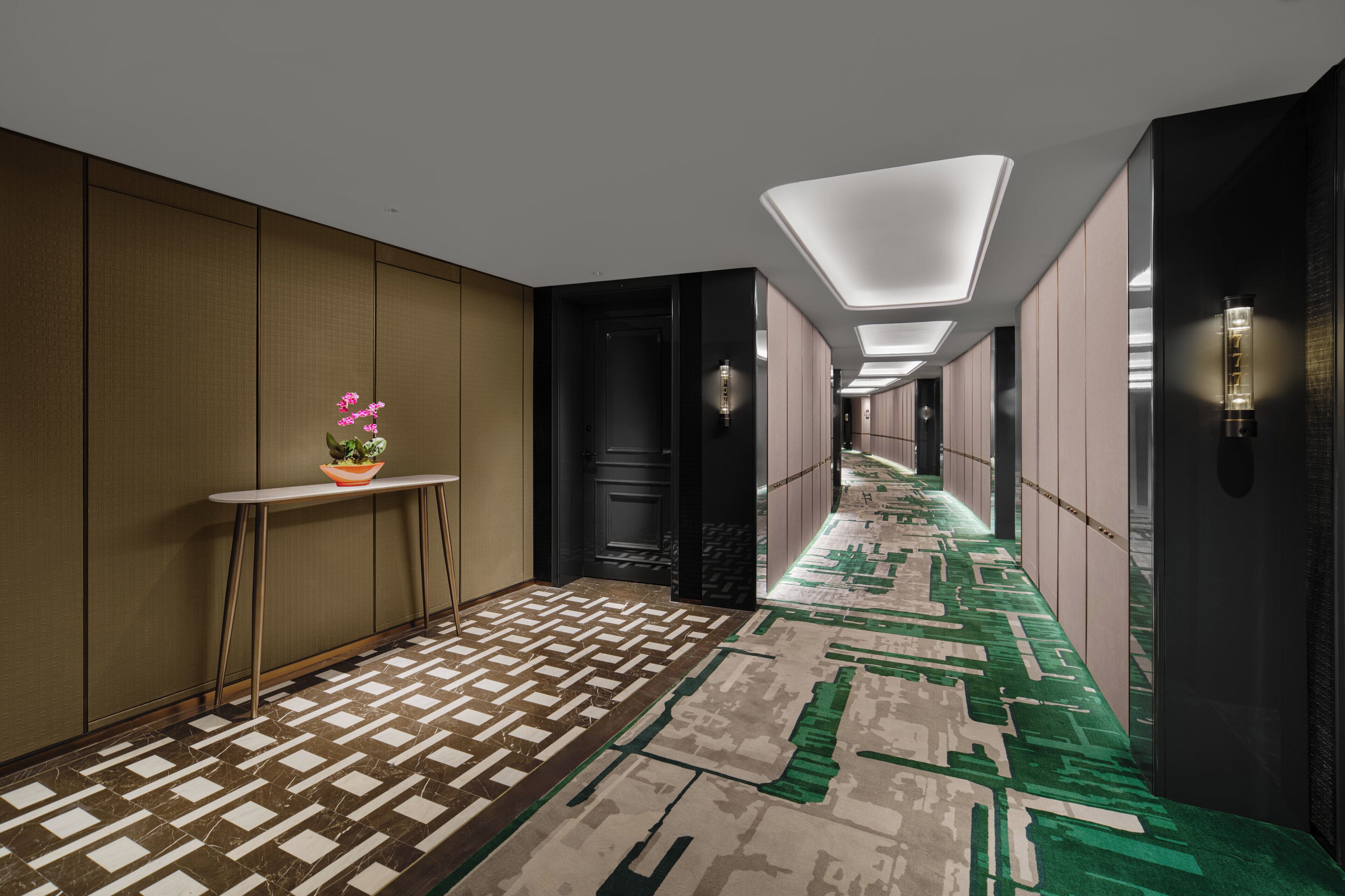 A modern hotel corridor with patterned flooring, a small table with a potted plant, and recessed lighting.
