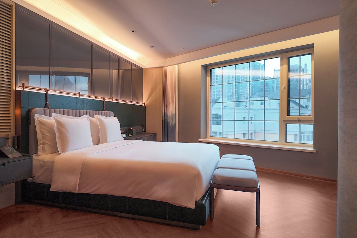 Un dormitorio moderno con una cama grande, armario empotrado, mesitas de noche y un amplio ventanal con vistas a la ciudad.
