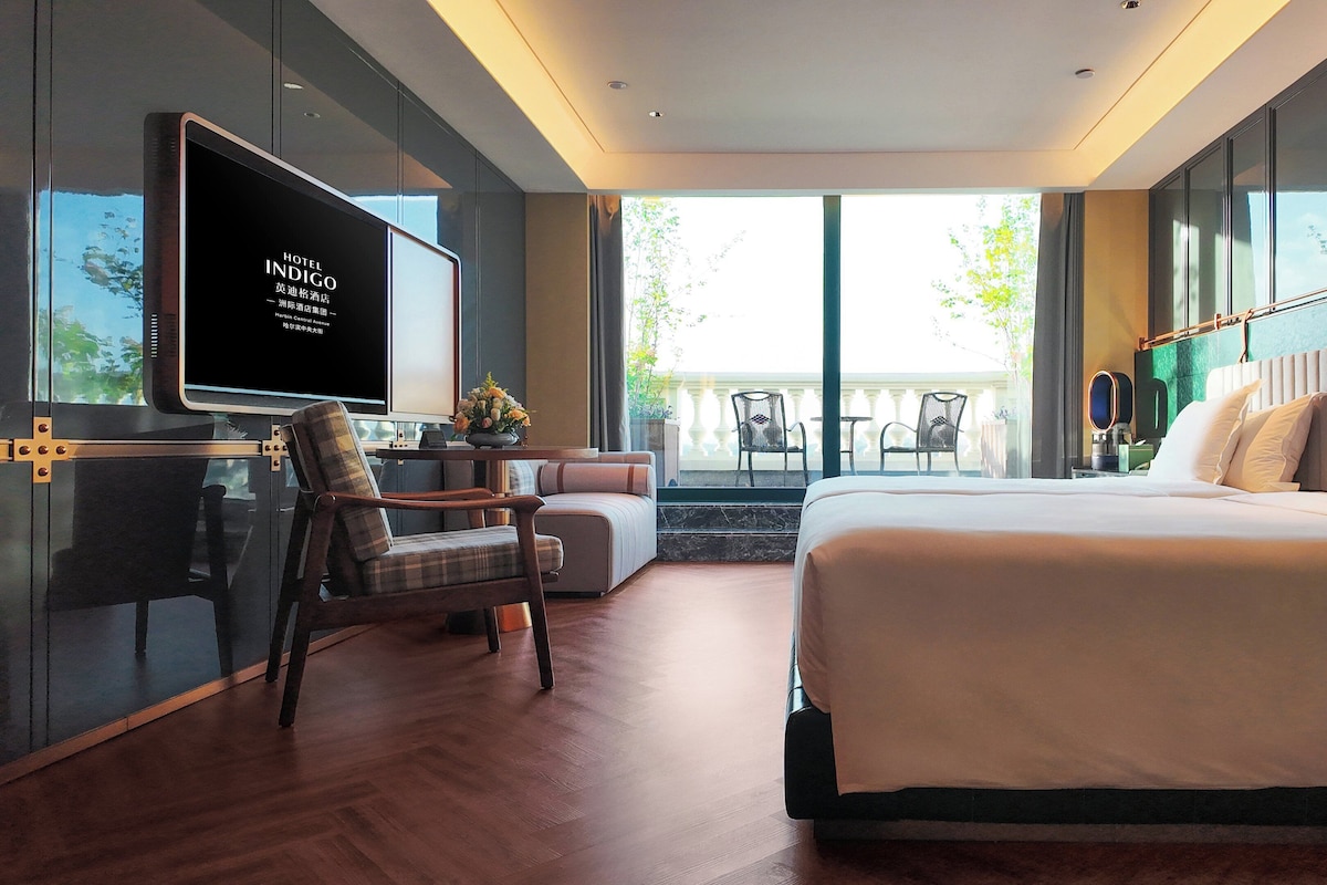 Una habitación de hotel moderna con una cama grande, un televisor, una zona de estar con una mesa y sillas, y un balcón con mobiliario exterior.