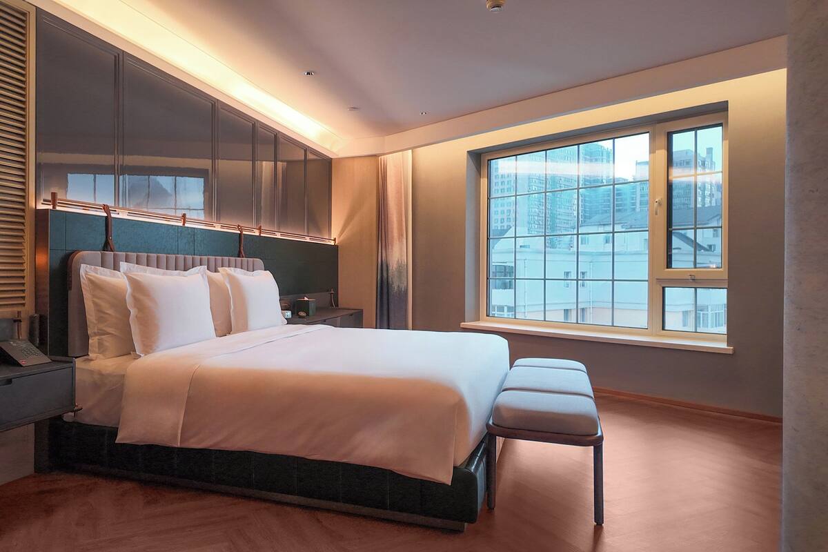 Une chambre moderne dotée d’un grand lit, d’une armoire intégrée, de tables de chevet et d’une grande fenêtre offrant une vue sur la ville.