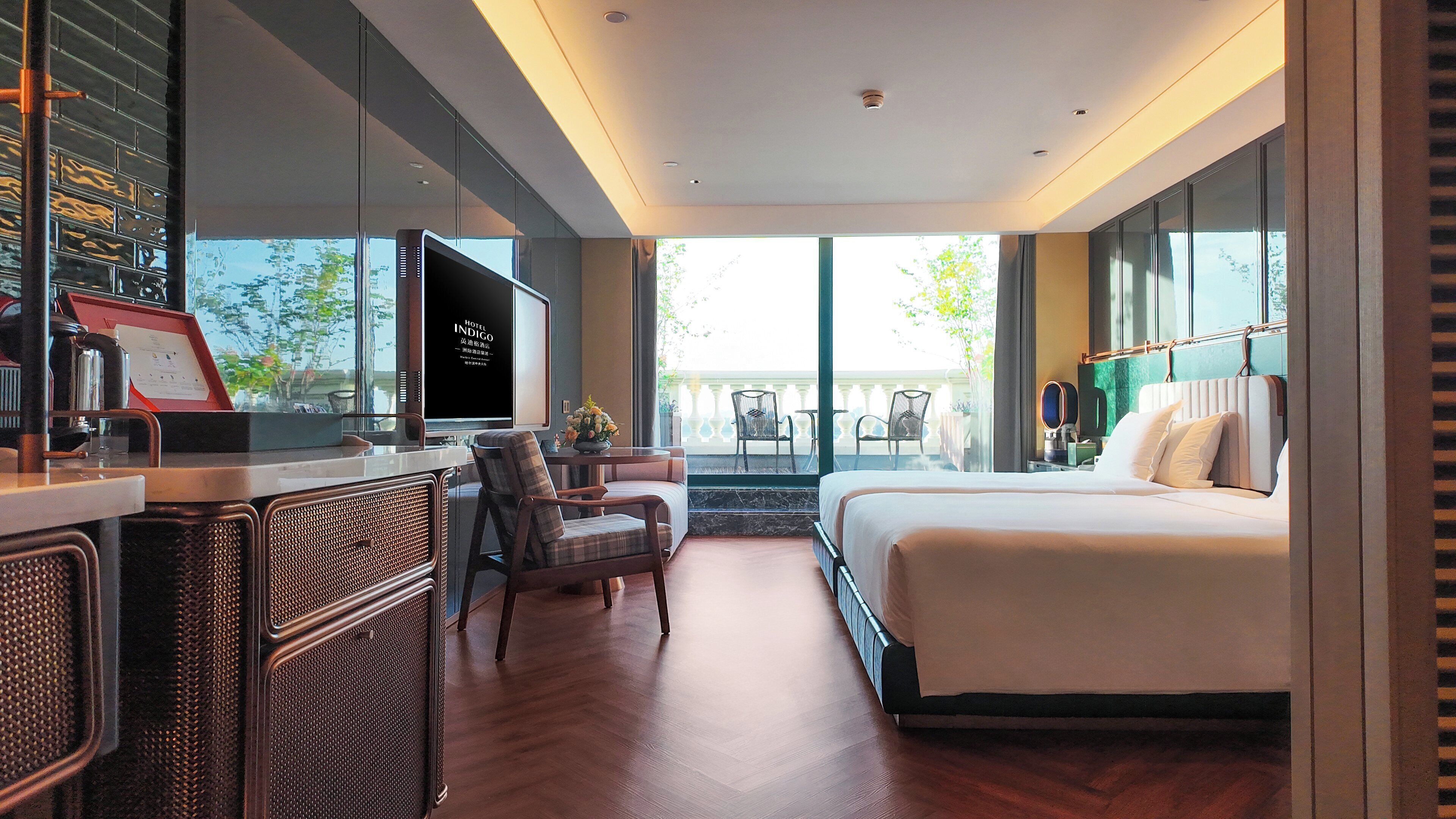 A modern hotel room with a large bed, a desk with a computer, a seating area with chairs, and a balcony with a view.