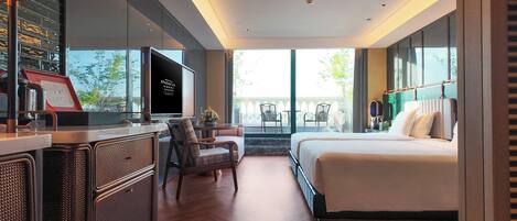 A modern hotel room with a large bed, a desk with a computer, a seating area with chairs, and a balcony with a view.
