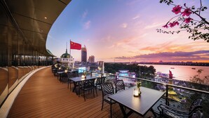 Una terraza en la azotea con mesas y sillas, vista del horizonte de una ciudad y un río con barcos.