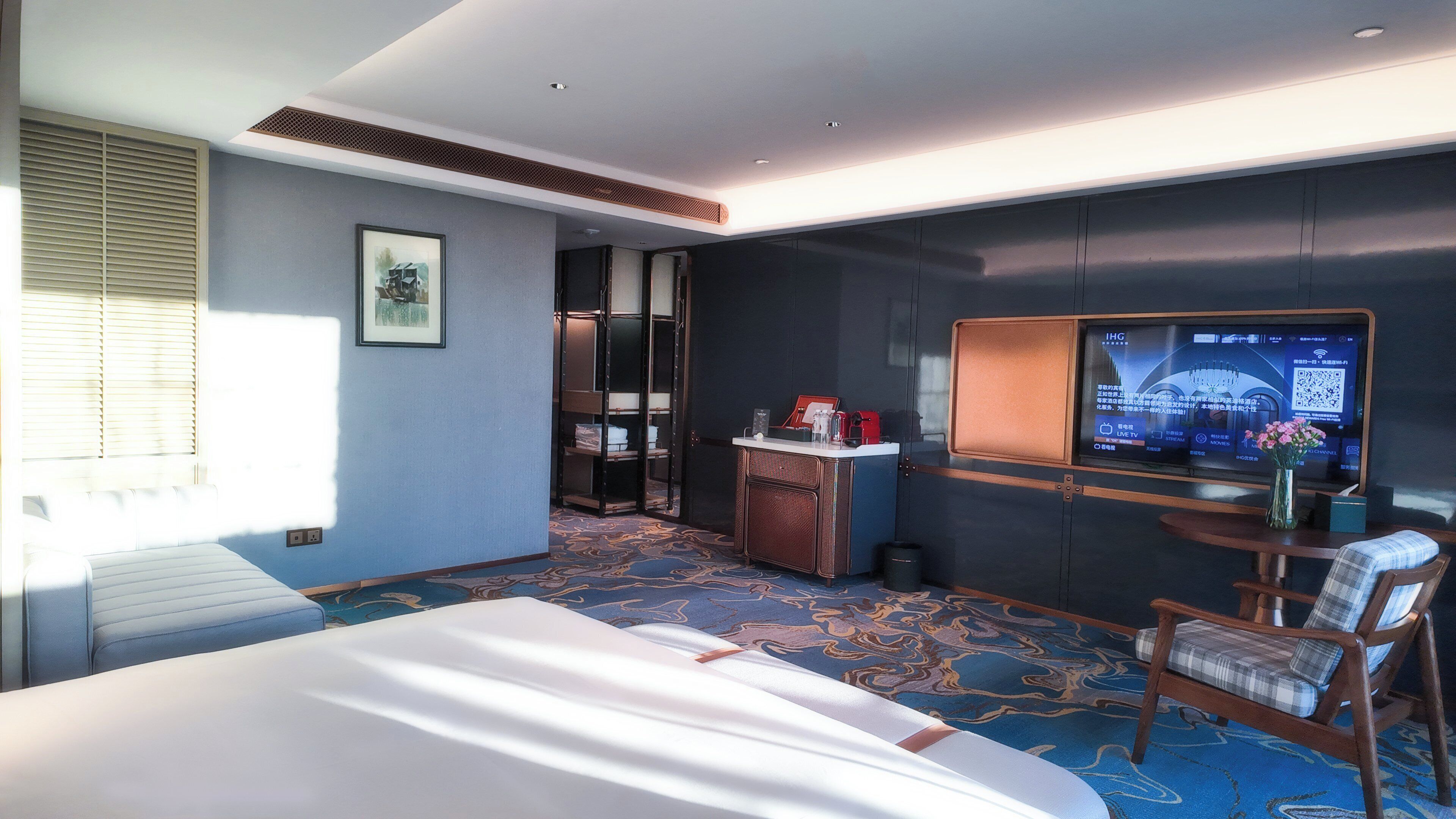 Quarto de hotel com cama, escrivaninha, computador, cadeira e uma janela grande com persianas.