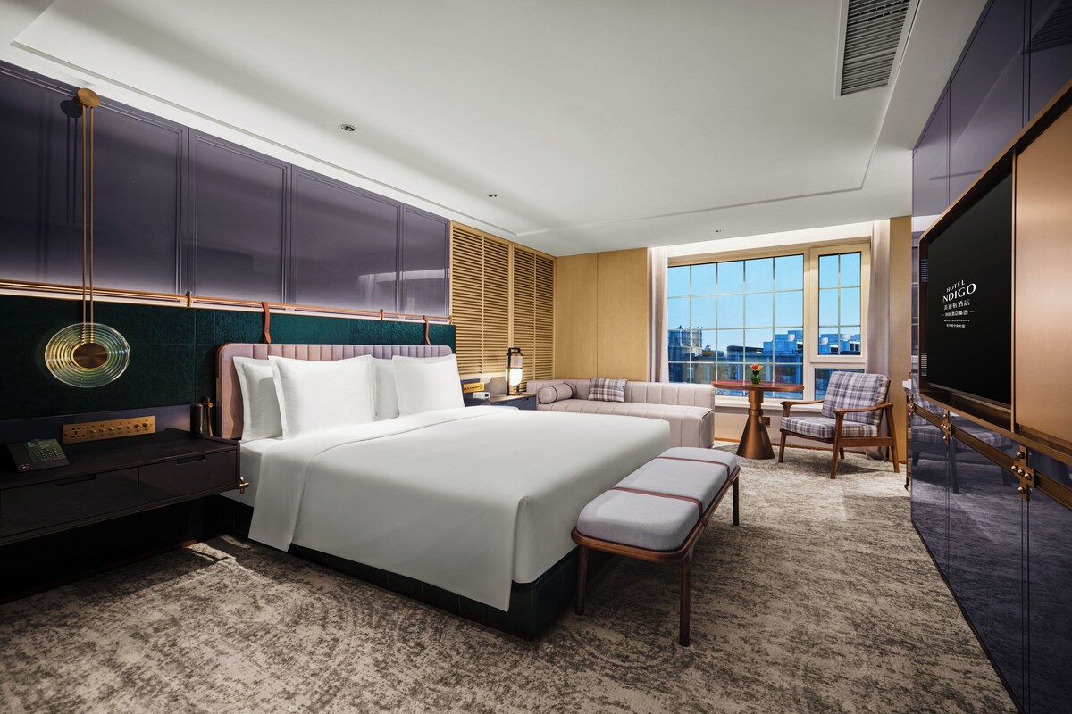 A modern hotel room with a large bed, a seating area with a sofa and chairs, a television, and a city view through the window.